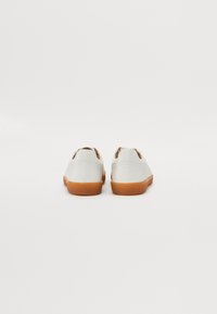 Chaussures en cuir blanc avec une finition texturée, semelles en caoutchouc marron et découpes au niveau du talon. Design minimaliste, sans marque visible.