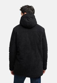 Zwarte corduroy jas met een capuchon, voorzien van een gestructureerd oppervlak en een naadloos achterontwerp, gedragen met donkere jeans.