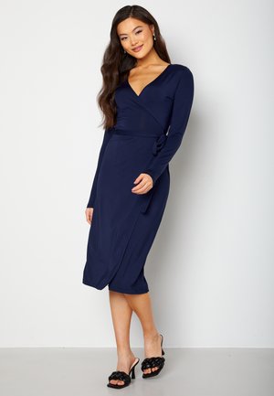 Abito avvolgente blu navy con maniche lunghe, scollatura a V e una cintura in vita. Abbinato a sandali neri con tacco caratterizzati da dettagli intrecciati.
