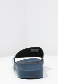 Bakifrån vy av en blå slide sandal med en strukturerad fotbotten, platt sula och ett svart rem. Enkel, modern design utan synliga mönster eller detaljer.