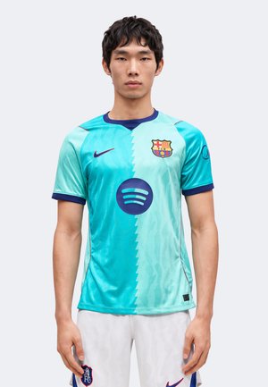 Jeune homme portant un maillot de football turquoise et bleu clair du FC Barcelone avec un short blanc, debout devant un fond uni.