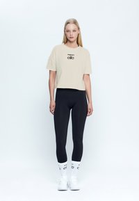 Camiseta recortada beige con logo negro, combinada con leggings negros, calcetines blancos con logo y zapatillas deportivas blancas. Diseño simple y ajustado.