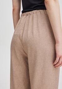 Ljusbruna linneblandade byxor med en texturerad yta, försedda med elastisk midja och sidofickor, visade från sidan.
