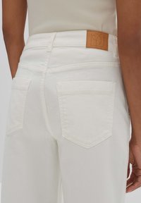 White denim jeans with a textured finish, feature two back pockets, and a tan leather patch on the waistband showing a minimalist logo.