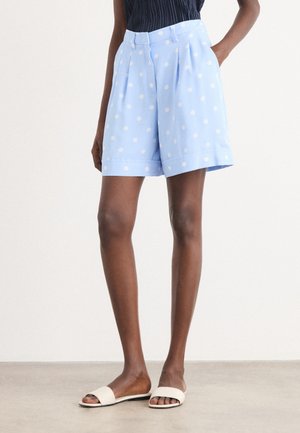 Woman wearing light blue high-waist shorts with white polka dots and white slide sandals, standing with one hand in pocket.