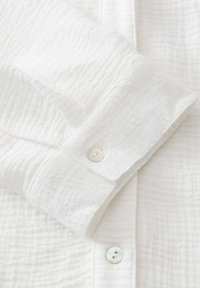 Poignet de chemise à manches longues texturé blanc avec deux boutons blancs sur un fond de tissu doux et froissé.
