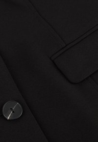 Zwarte wollen mix jas met een gladde textuur, strakke lijnen, een omgeslagen kraag en een ronde knoop met twee gaatjes als sluiting.