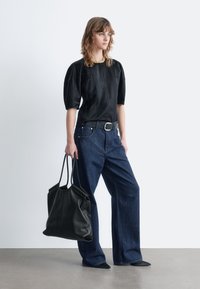 Woman wearing a black puff-sleeve top, blue wide-leg jeans, black belt, pointed shoes, gold necklace, holding a large black tote bag.