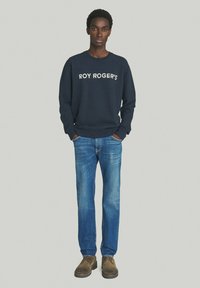 Felpa navy con la scritta "ROY ROGER'S" in lettere bianche, abbinata a jeans blu medio e scarpe marroni. Vestibilità casual, polsini e orlo a costine.