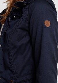 Marineblaue Jacke aus robustem Stoff mit Reißverschluss und Druckknopfverschluss, mit einem braunen Leder-Logoaufnäher auf der linken Schulter.
