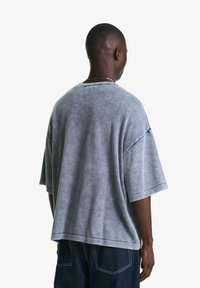 Šedé oversize tričko s pokleslými rameny, měkkou texturou a surovým lemem. Zadní pohled ukazuje minimalistický design a volný střih.