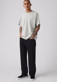 Bărbat cu brațe tatuate, purtând un tricou Levi's gri deschis, pantaloni negri și pantofi slip-on negri, stând cu mâinile în buzunare.