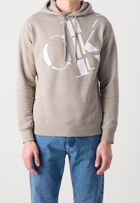 Sudadera beige con un bolsillo tipo canguro, estampado gráfico blanco en el frente y cordones. Tejido texturizado y puños y dobladillo acanalados.