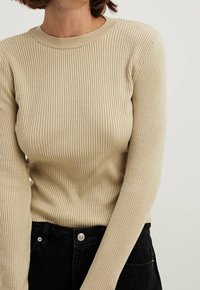 Haut en tricot beige à manches longues côtelé avec un col rond. Il présente des manches ajustées et une texture lisse, associé à un pantalon noir taille haute.