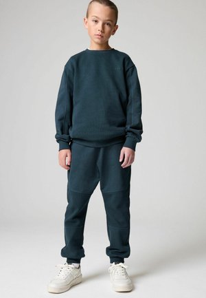 Tumman teal-värinen huppari, jossa on teksturoitu kuvio, yhdistettynä samansävyisiin verkkareihin, joissa on paneelikorostuksia; viimeistelty valkoisilla lenkkareilla.