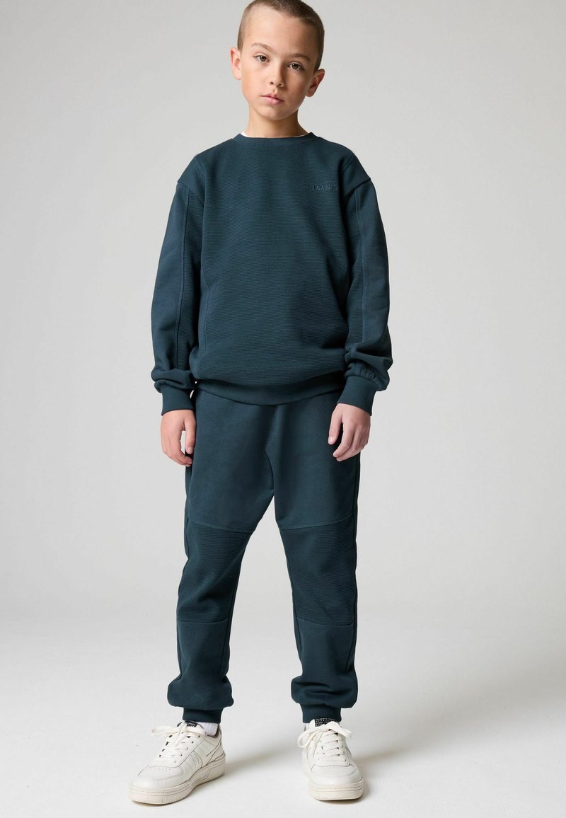 Tamsiai žaliai mėlyna džemperis su tekstūriniais raštais, derinamas su atitinkamomis sportinėmis kelnėmis, turinčiomis panelių akcentus; užbaigtas baltais sportbačiais.