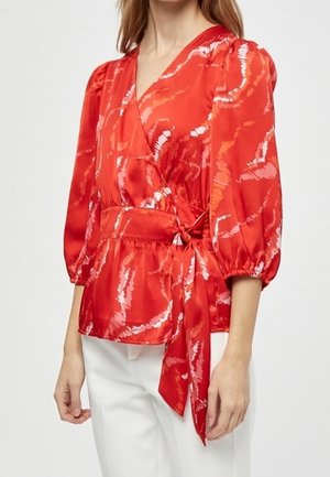Femme portant un chemisier portefeuille en satin rouge avec des motifs abstraits et des manches trois-quarts bouffantes, associé à un pantalon blanc.