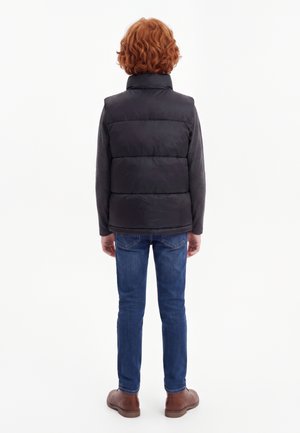 Enfant aux cheveux roux bouclés portant un gilet matelassé noir, un t-shirt à manches longues sombre, un jean bleu et des chaussures marron, debout de dos.