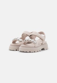 Sandales beige clair avec de larges sangles et un accent décoratif en strass sur le bout. Dotées d'une semelle épaisse et texturée pour une meilleure adhérence.
