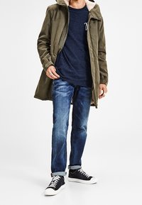 Olivově zelená parka s kapucí zdobenou kožešinou, námořnické tričko, modré džíny a černé tenisky s bílou podrážkou. Neformální městský outfit.