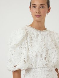 Λευκό τοπ από δαντέλα με περίπλοκα λουλουδάτα σχέδια, φουσκωτά μανίκια και λαιμόκοψη διακοσμημένη με πέρλες. Λεπτός και υφασμένος σχεδιασμός.