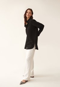 Langarmhemden in Schwarz mit Knöpfen, weiße weite Hosen und spitze flache Schuhe mit schwarz-weißem Muster. Glattes Material mit Seitenschlitzen.