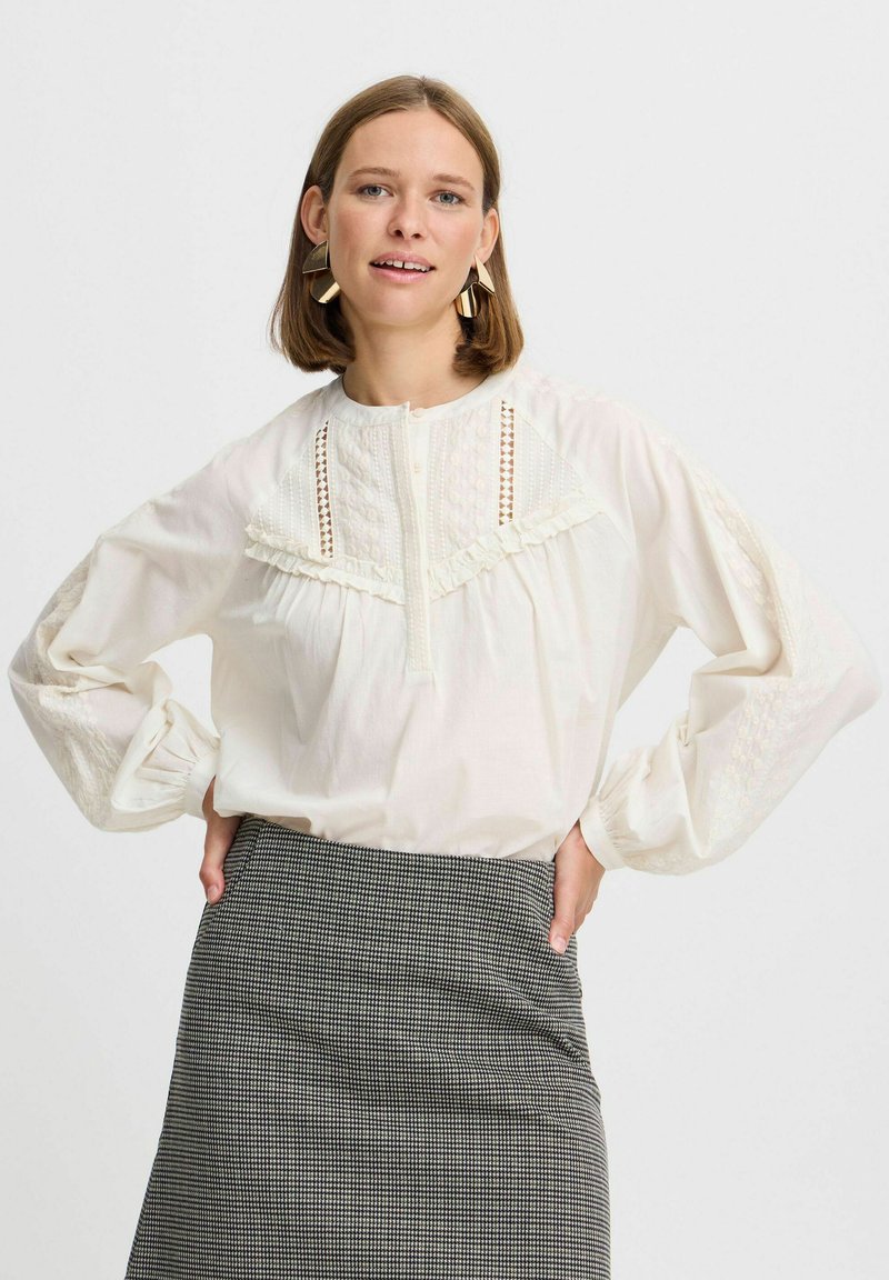 Weiße Bluse mit filigraner Spitze und Rüschendetails, Knopfverschluss am Hals, lange Ärmel mit strukturierten Akzenten. Kombiniert mit einem gemusterten Rock.