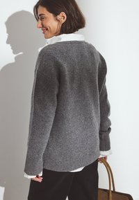 Grauer Strickpullover mit geripptem Saum und Bündchen, über einem weißen Hemd getragen, gehalten mit einer braunen Handtasche. Hat einen lockeren Schnitt und eine weiche Textur.