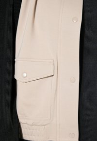 Beige leichte Jacke mit einer geknöpften Klappentasche und glatter Textur, kombiniert mit einem schwarzen gerippten Stoff darunter.