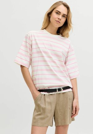 Blond kvinne iført hvit oversized T-skjorte med rosa striper, beige shorts og svart belte, stående mot ensfarget lys bakgrunn.