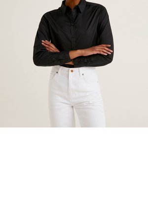 Personne portant une chemise noire à manches longues boutonnée et un pantalon blanc taille haute, les bras croisés au niveau de la taille.