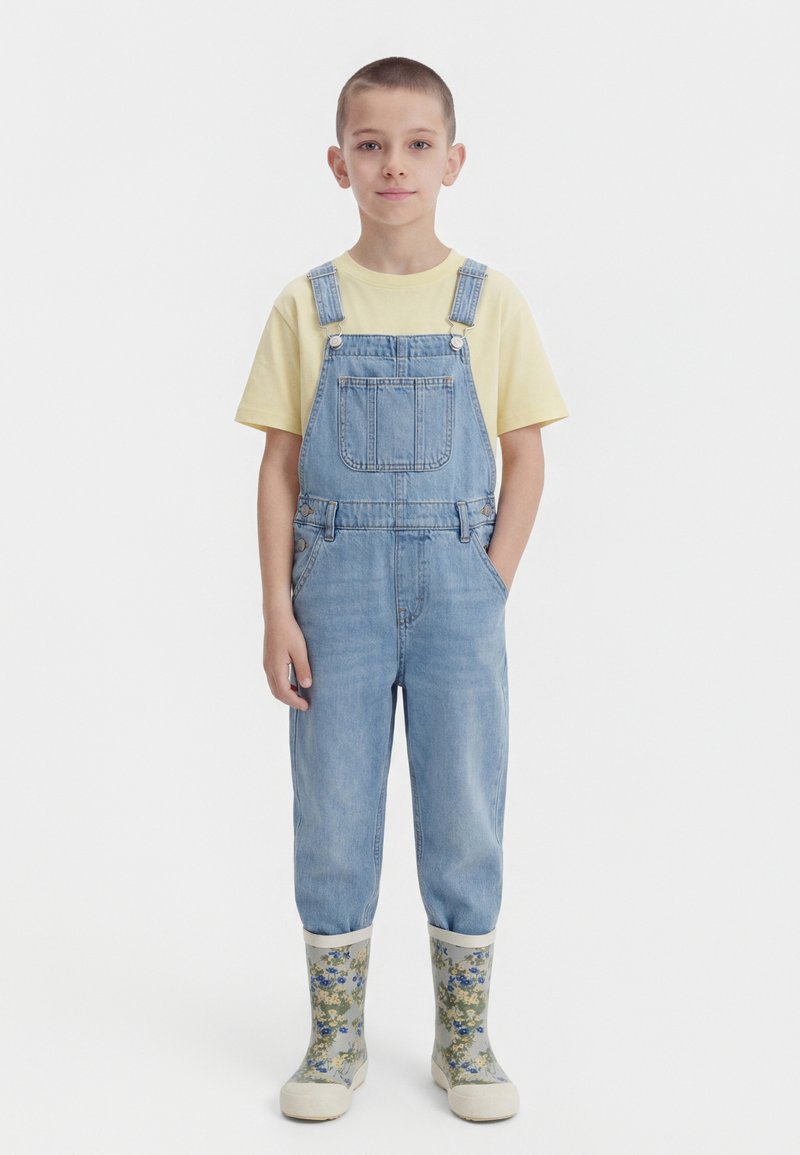 Băiat tânăr, stând cu mâinile în buzunare, purtând un tricou galben, salopetă de denim albastru deschis și cizme de ploaie cu model floral.
