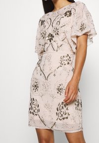 Pärlad klänning i ljus rosa, med en lös design med fladderärmar, prydd med intrikata blommönster och paljettdekorationer genom tyget.