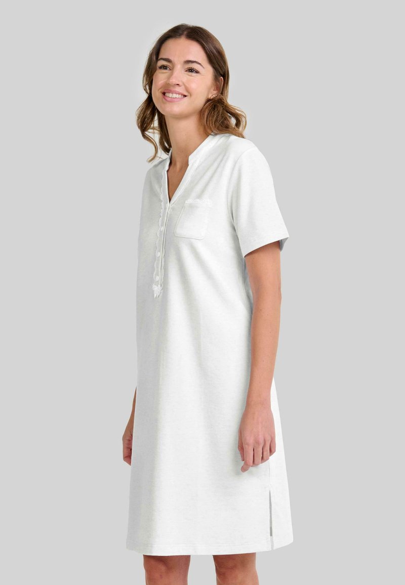 Mujer sonriente vestida con un vestido blanco de manga corta y hasta la rodilla, con un bolsillo frontal y un escote en V abotonado, de pie contra un fondo gris claro.