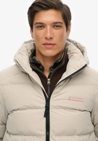 Chaqueta de plumas beige con capucha, con un zipper negro, textura acolchada y un logo rojo en el pecho. Lleva una capa sobre un suéter marrón.