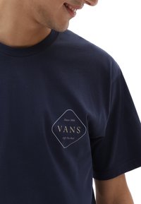 Tummansininen puuvillapaita, jossa on timantin muotoinen logo, jossa on "VANS" kultaisella kirjoituksella ja teksti "Since 1966" sen yläpuolella. Lyhyet hihat.