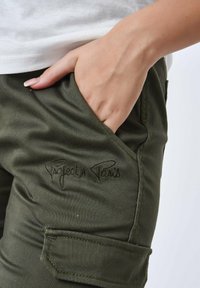 Pantalones cargo de tela verde con un bolsillo frontal y el logo bordado "Project x Paris". Las características incluyen una textura suave y detalles de costura.