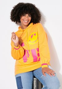 Felpa gialla brillante con una grafica di un sorriso rosa, maniche arricciate e una vestibilità comoda. Abbinata a jeans con dettagli in patchwork.