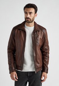 Brun læderjakke med kviltede skulderdetaljer, lynlås foran og brystlomme. Båret over en lys grå sweater, parret med mørke jeans.