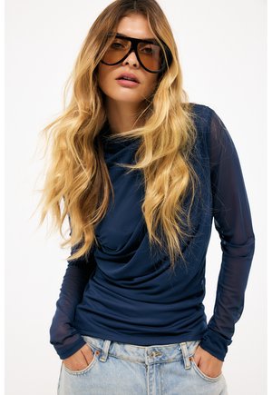 Femme aux longs cheveux blonds portant des lunettes de soleil surdimensionnées teintées ambré, un haut à manches longues drapé bleu marine et un jean bleu clair, les mains dans les poches.