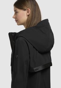 Veste à capuche noire en tissu léger, dotée d'une fermeture éclair, de poches latérales et d'une sangle avec logo au dos.