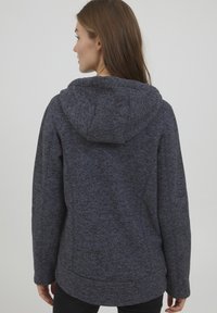 Veste à capuche en polaire texturée gris foncé, avec une coupe décontractée, présentant un ourlet arrondi et des détails de couture subtils sur les manches.