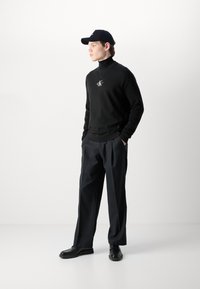 Black turtleneck sweater with ribbed cuffs and a small logo, paired with loose-fitting gray trousers and black shoes, complemented by a black cap.