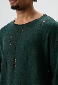 Groene gebreide trui met een brede halslijn, voorzien van meerdere versleten gaten en een gestructureerde stof. De randen zijn onafgewerkt en lichtjes rafelig.