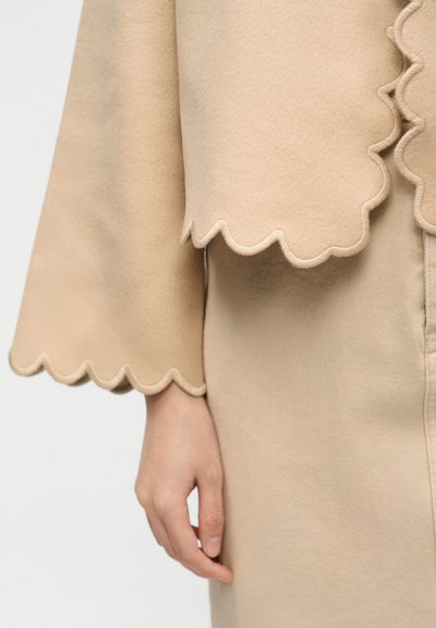 Beige mouw van een jas en rok met gegolfde randen, gedeeltelijk arm en hand zichtbaar tegen een effen achtergrond.