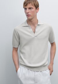 Ung mand iført en lysebeige kortærmet strikket polo og hvide bukser, stående foran en ensfarvet lys grå baggrund, med hænderne i lommerne.