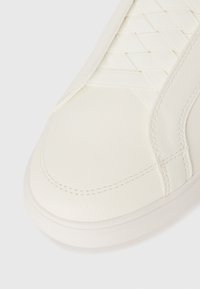 Acercamiento del dedo del pie de una zapatilla blanca que presenta cuero texturizado, detalle de correa diagonal y costuras visibles en la suela y la parte superior.