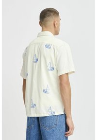 Kurzarmhemd aus cremefarbenem Stoff mit blauer Segelboot-Stickerei. Lockerer Schnitt mit Kragen und geradem Saum. Leichte Textur.