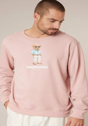 Man draagt een lichtroze sweatshirt met een illustratie van een beer met zonnebril, een blauw overhemd, witte broek en sandalen op de borst.