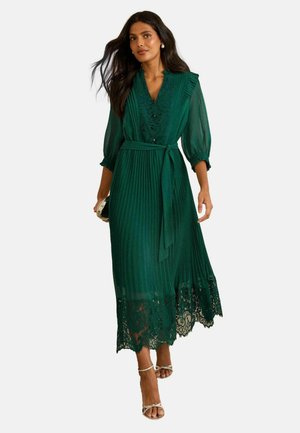 Grünes plissiertes Kleid mit V-Ausschnitt, 3/4-Ärmeln, Spitzenbesatz und einem Stoffgürtel. Getragen mit silbernen Riemchenschuhen und einer Clutch.